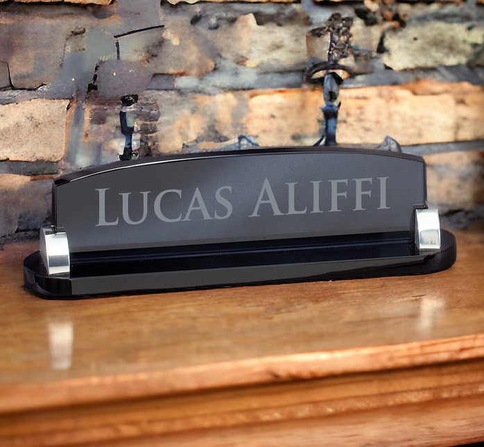 Our smoked glass desk name plate. It features silver metal holders on each side. The name plate is laser engraved with the recipients name. It's sitting on a wood desk with a brick background.