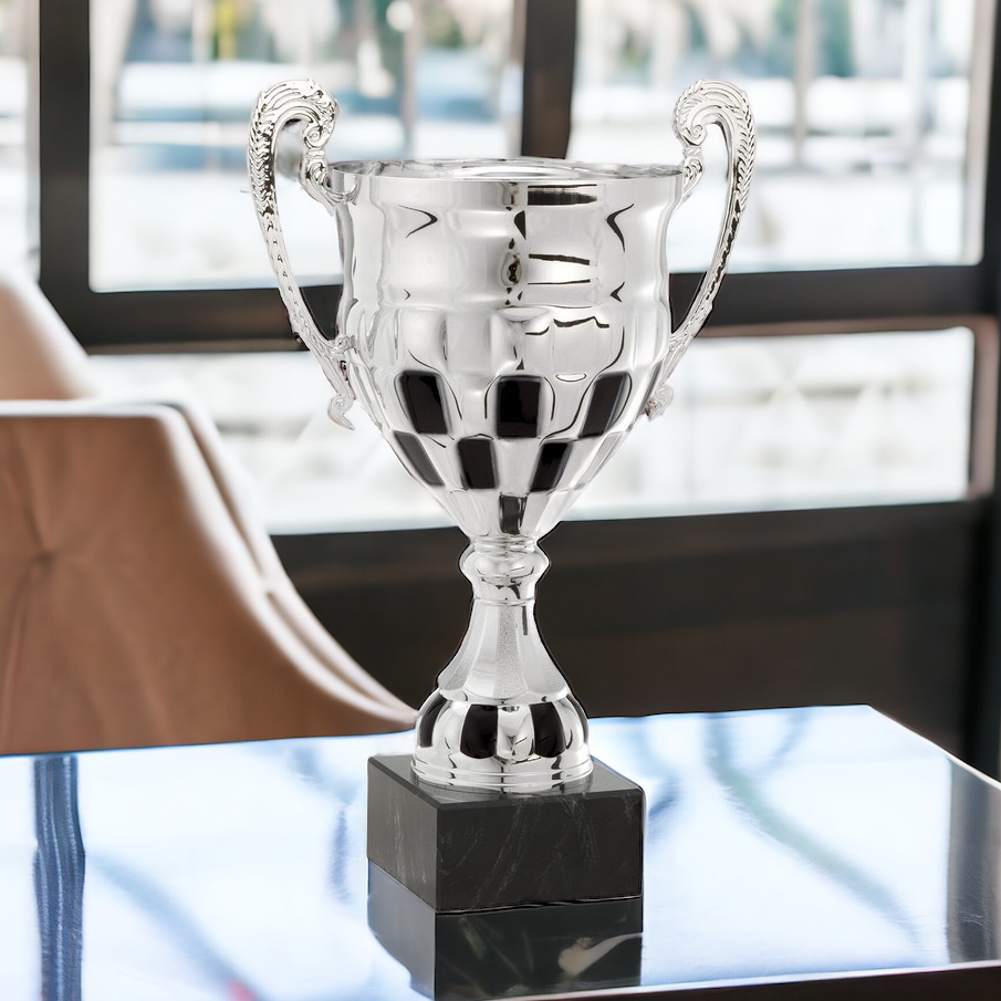 A racing trophy cup made with silver metal. It features two handles, a checkered flag design and is mounted on a black marble base. This trophy cup is sitting on a table for display with a window in the background.