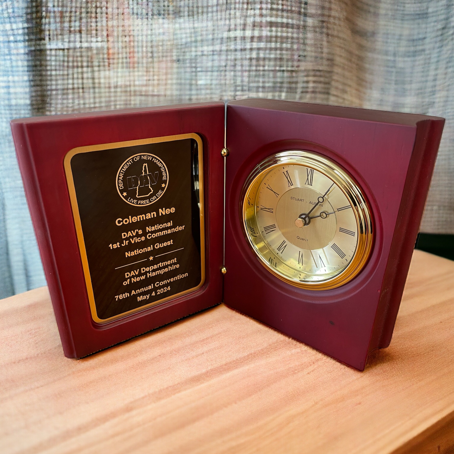 A book clock made with a hand-rubbed mahogany finish. The inside features a black & gold engraving plate on the left and a gold clock with a lifetime warranty on the right. It's sitting on a desk with curtains in the back ground.