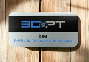 A silver name badge that is imprinted with full color. The company's logo is 3DPT and the name badge is made for Kim who is a Physical Therapist Assistant.