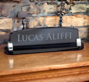 Our smoked glass desk name plate. It features silver metal holders on each side. The name plate is laser engraved with the recipients name. It's sitting on a wood desk with a brick background.