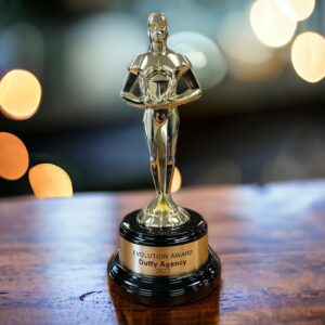 Our Classic Achiever Statue resembles the classic Oscar Statue. It features a man holding a wreath made from heavy metal & coated in a glossy gold color. It's mounted on a glossy black base that includes a gold engraving plate for personalization. This statue is sitting on a table with lights in the background.