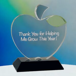Our Teacher Apple features a clear crystal apple with a green leaf accent at the top. The middle area is for personalization and this specific apple says, "Thank you for helping me grow this year!" It's mounted on a black crystal base. This crystal apple is sitting on a white table with a blue & green background.