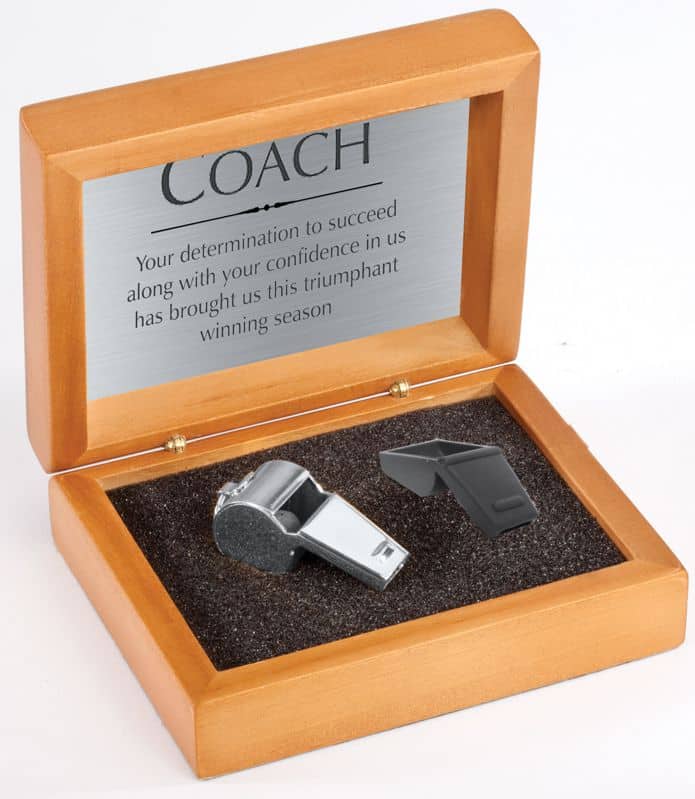 A wood box with a silver engraving plate on the inside lid that is personalized with an appreciative message for a coach. Below is a silver whistle on the left and a whistle cover on the right.