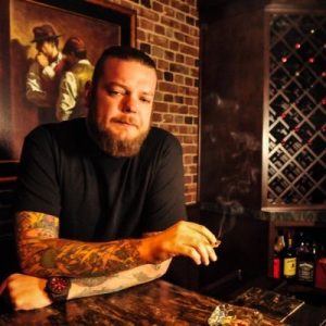 A photo of Cory Harrison of the TV Show Pawns Stars. He's sitting at a bar with a cigar in his hand. He has a clean shaven beard, slicked back hair, tattoos on his arms and a watch on his left hand.