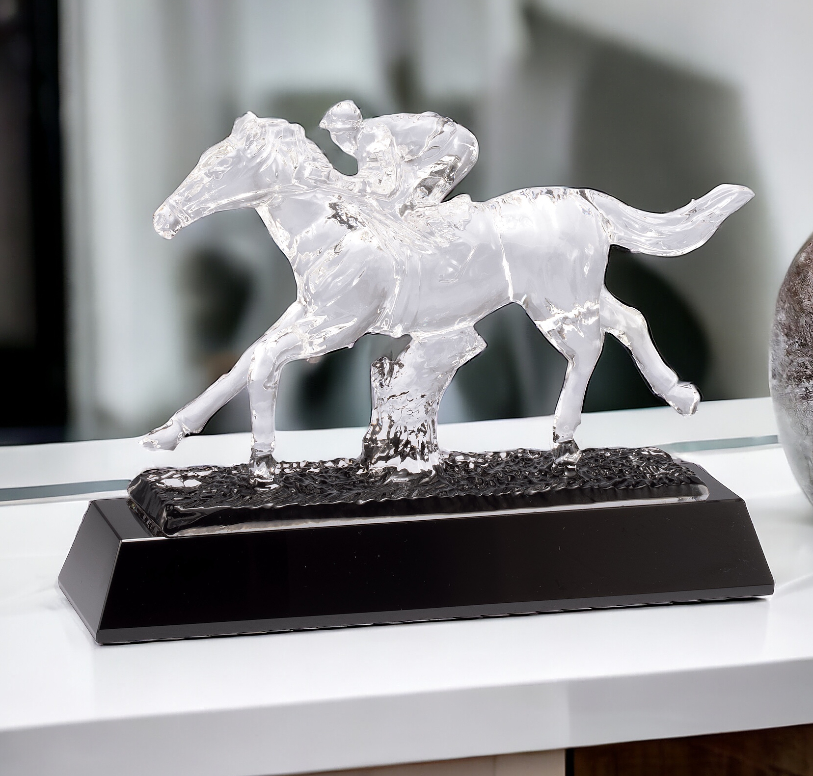 Our Crystal Horse Racing Trophy sitting on a white shelf. The trophy features a clear crystal horse & jockey mounted on a black crystal base. The base includes a black & silver engraving plate for personalization. This CRY599 Horse Racing Sculpture is 7" x 9.5" in size and weighs over 5 lbs.