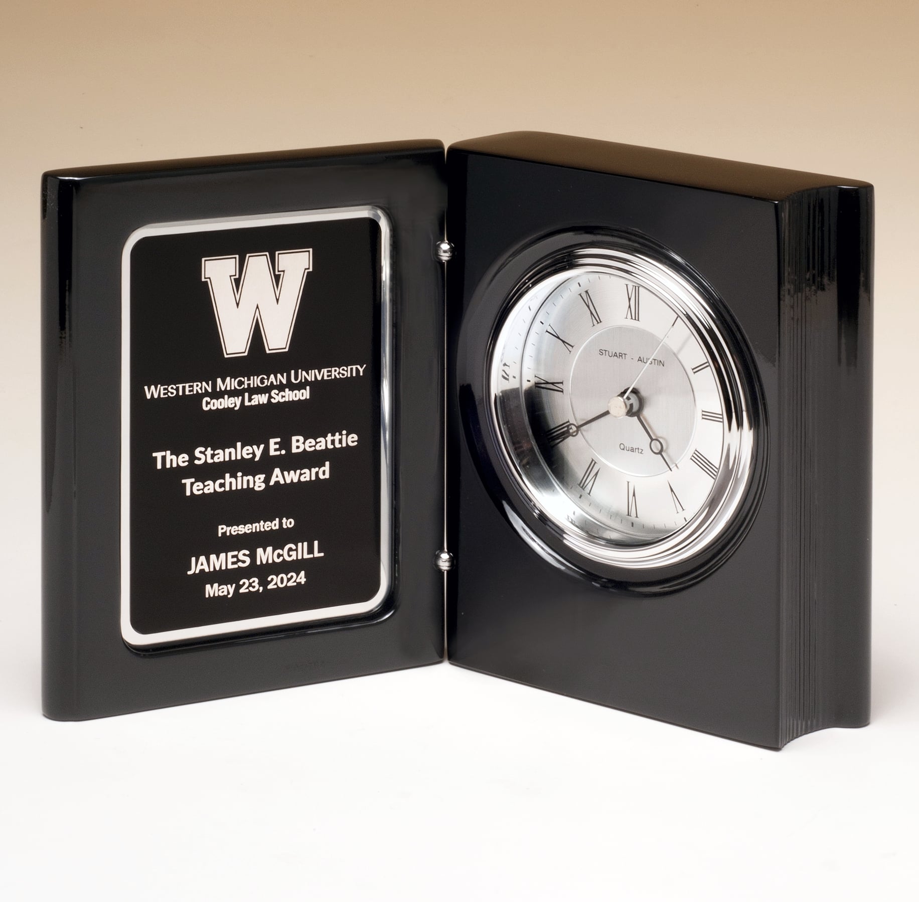 A black book clock that features a piano finish on the wood. On the inside is a silver faced clock & bezel with black numerals on the right and a black & silver engraving plate on the left.