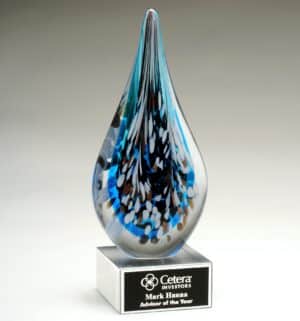 An art glass award in the shape of a rain drop with black, white & gray colors speckled in front of the predominant blue color. It's mounted on a clear glass base that includes a black & silver engraving plate for personalization.