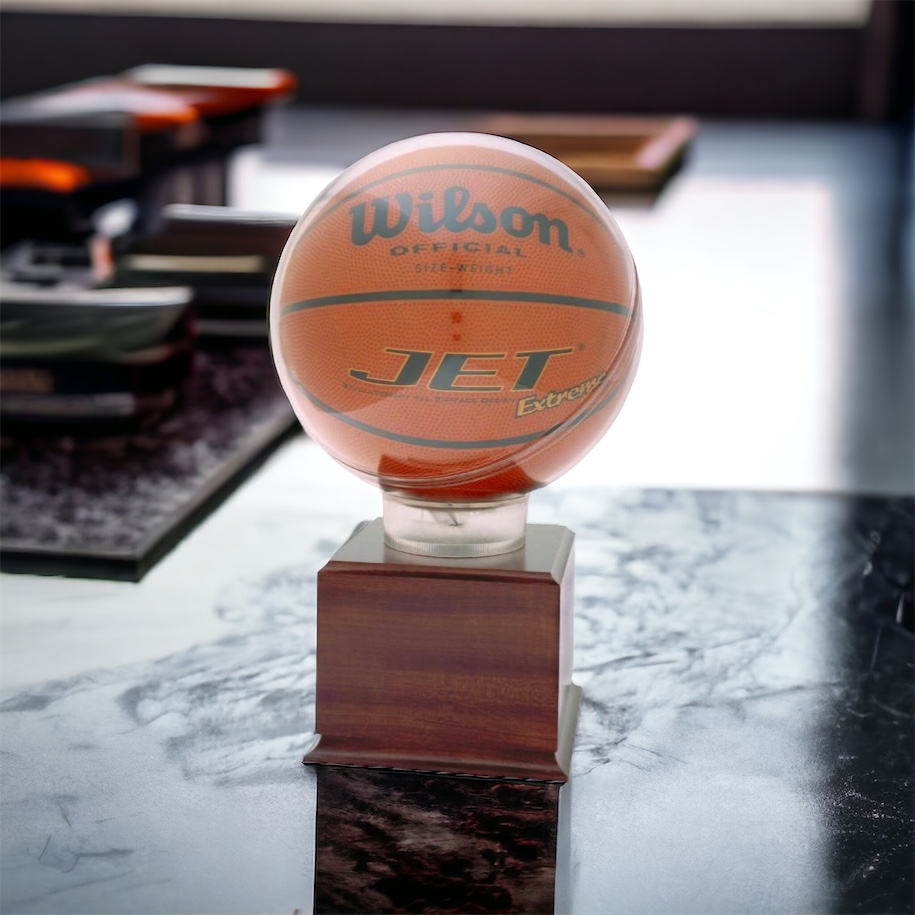 A basketball display case with a round acrylic area to hold the ball. It's mounted on a larger wooden base that includes an engraving plate for personalization. The display case is sitting on a marble desk.