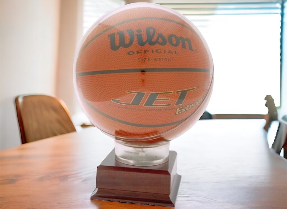 A basketball display case featuring a round acrylic holder for the ball that is mounted on a wooden base. The base includes an engraving plate for personalization. The basketball display case is sitting on a table with chairs around it and a window in the background.
