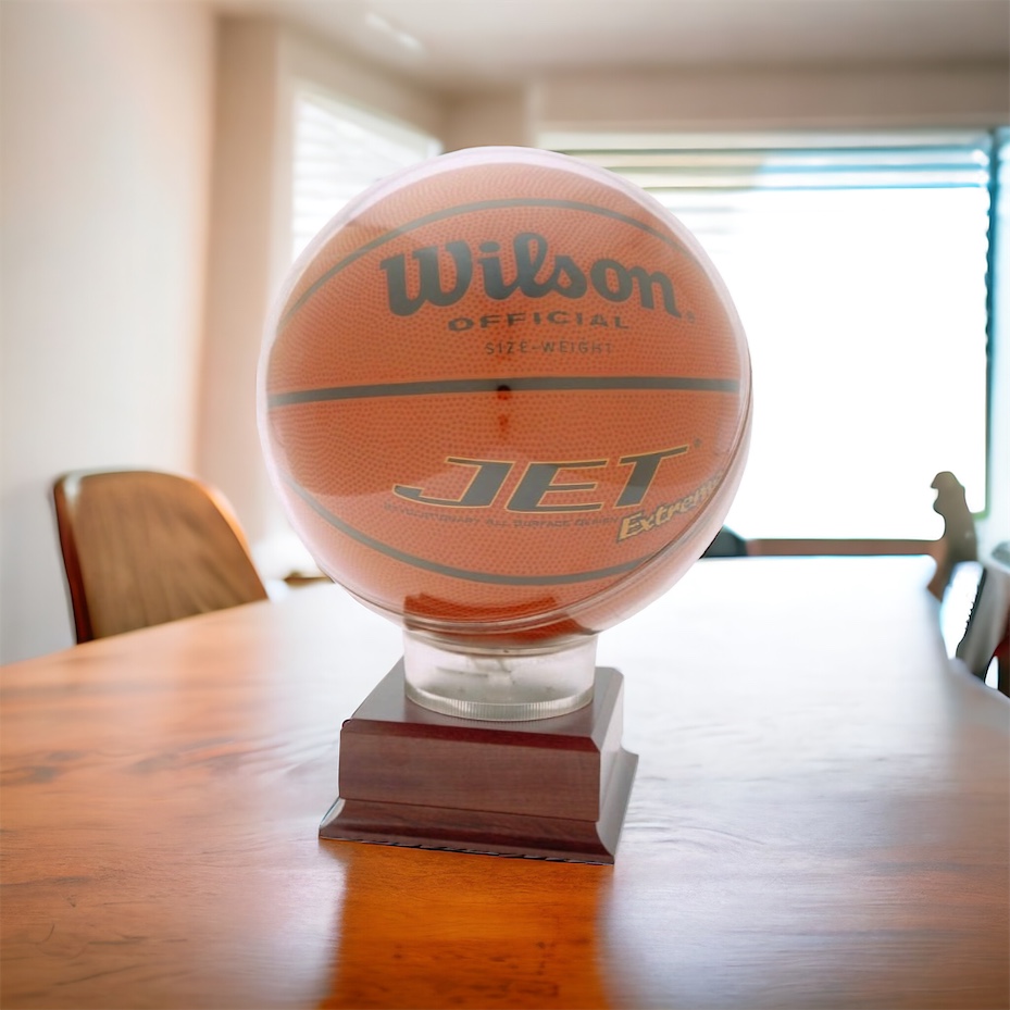 A basketball display case featuring a round acrylic holder for the ball that is mounted on a wooden base. The base includes an engraving plate for personalization. The basketball display case is sitting on a table with chairs around it and a window in the background.