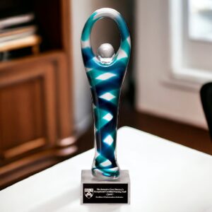 A Victory Glass Art Award featuring a contemporary human with their arms raised above their head & holding their hands at the top. For added decoration there are teal & navy colors throughout the body. It's mounted on a clear glass base with a black & silver engraving plate for personalization. This specific award has been personalized for The Intensive Care Nursery's Exceptional Certified Nursing Staff.