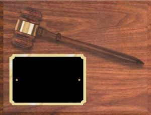 A walnut finished board with a walnut gavel & gold gavel band. The gavel slants from top left to middle right. Below the hammer of the gavel is a black engraving plate with notched corners & a gold border.