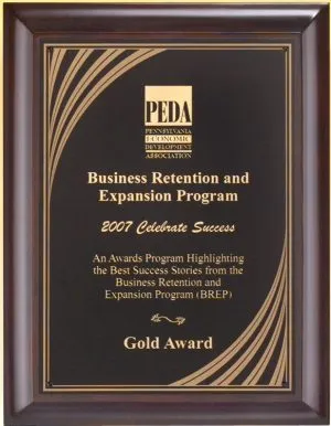 A cherry wood board with a black & gold engraving plate featuring a cascade design in the corners. The middle of the engraving plate features a company logo and words of recognition for an employee.