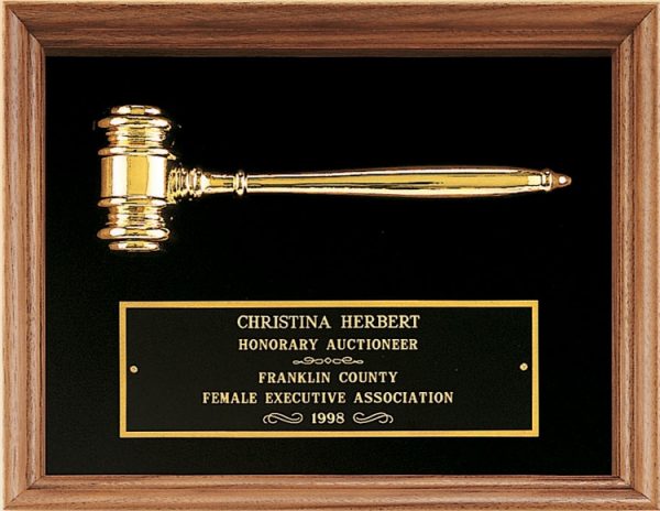 A walnut frame with black velour on the inside. At the top is a gold metal gavel. At the bottom is a black & gold engraving plate for an honorary auctioneer.