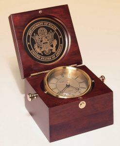 A Captain's Clock made with a mahogany finish. The lid has a round, black engraving plate with the Department of State logo laser engraved into it. The bottom of the box features a gold clock that can swivel with the knobs that stick out the sides.