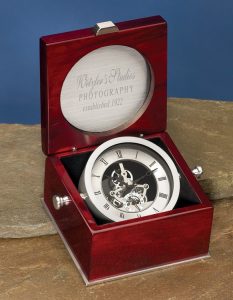 A captain's clock made with a rosewood box with a silver engraving plate in the top for personalization. The bottom of the box features a silver skeleton clock that can be swiveled with the handles on each side.
