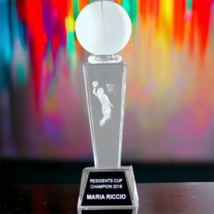 Women's Basketball Trophy made entirely of crystal. It features a small, solid crystal basketball at the top, the middle area has a 3D version of a female attempting a dunk & a base the includes an engraving plate for personalization. The trophy is on a desk and the background of the phot is very colorful.