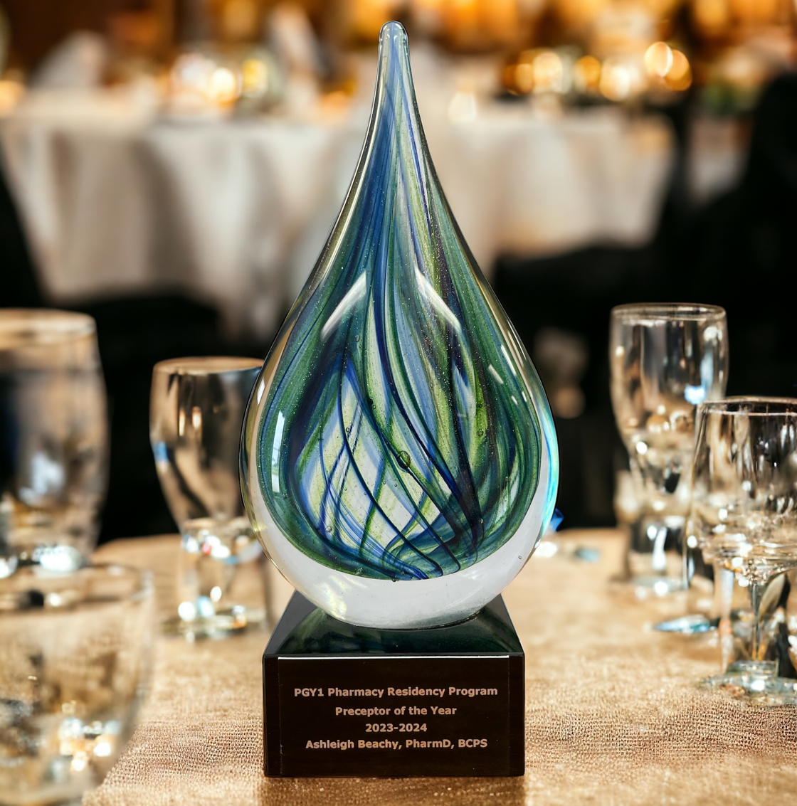 Our Green Teardrop Art Glass Award features a solid glass teardrop with green & metallic gold throughout. It's mounted on a black glass base with a black & gold engraving plate for personalization. This one is sitting on a gold table at an awards ceremony.