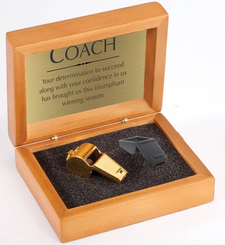 A wood box with a black bottom holding a gold whistle and a black whistle cover. The top of the box features a gold engraving plate with the words of appreciation towards a coach.