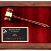 A smooth walnut wood frame with rounded corners. Inside the frame is a maroon velour background. On the velour is a walnut gavel with a gold gavel band at the top. In the bottom left hand corner is a black & gold engraving plate for personalization. The engraving features a company logo and words of recognition for a Chairman.