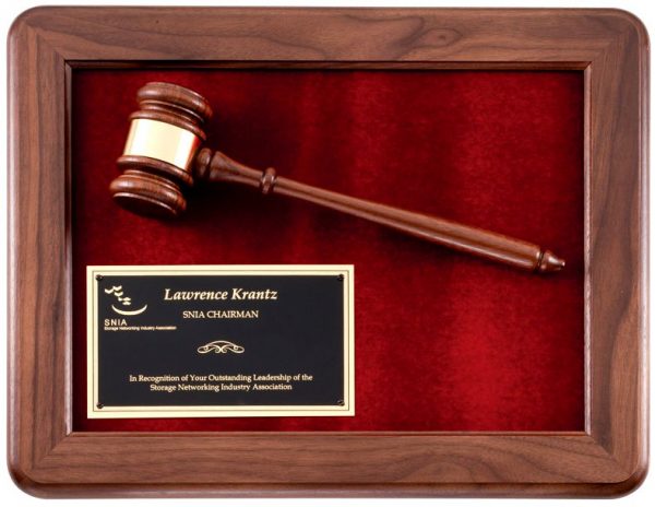 A smooth walnut wood frame with rounded corners. Inside the frame is a maroon velour background. On the velour is a walnut gavel with a gold gavel band at the top. In the bottom left hand corner is a black & gold engraving plate for personalization. The engraving features a company logo and words of recognition for a Chairman.