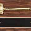 A walnut finish board with a gold metal gavel laying horizontally across the top and a black engraving plate towards the bottom of the plaque.