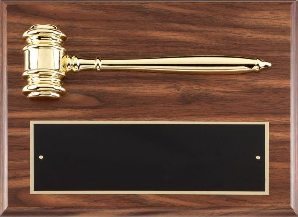 A walnut finish board with a gold metal gavel laying horizontally across the top and a black engraving plate towards the bottom of the plaque.