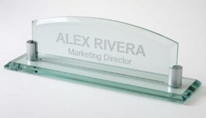 Deluxe glass desk name plate featuring a thick, crystal-clear curved glass panel with laser-engraved personalization. The name 'ALEX RIVERA' appears prominently in large frosted lettering, with 'Marketing Director' in smaller text below it. The elegant name plate is mounted on a beveled glass base with two sturdy silver aluminum supports, shown against a clean white background.