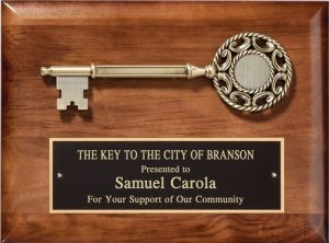 A 9x12 walnut board with a cast a cast metal, large decorative key at the top. Below is a black brass engraving plate for personalization. The engraving is for The Key to the City of Branson and it's presented to Samuel Carola.