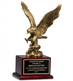 Antique Bronze Eagle Statue (1588) featuring a large, highly detailed resin eagle in a dramatic mid-strike pose with powerful outstretched wings, finished in a rich antique bronze glaze. The eagle is mounted on a luxurious piano-finish rosewood base with a black and gold engraving plate that reads: 'LEADERSHIP AWARD / Presented To MARCUS RITTER / For Your Dedicated Service and Commitment to Excellence / 2017'. This impressive eagle statue is 9.5″ wingspan x 15″ tall (7 lbs) trophy