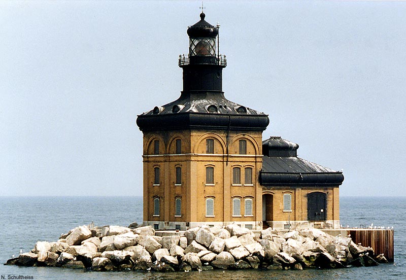 A tan colored lighthouse with a black roof. It appears to be build on an a man made island with large boulders surrounding it's base.