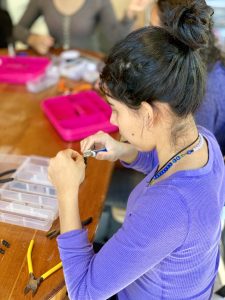 A photo of a woman at El Refugio in Guatemala using pliers to to create jewelry. She's wearing a purple shirt and on the table is a pliers & other things to make jewelry.  