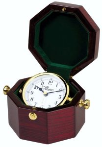 A captain's clock made with piano finished rosewood in an octagon shaped box. The inside is lined with green felt and features a white face clock with a gold bezel. The clock can swivel with the use of the gold knobs on the outside of the box.