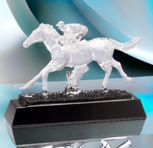 A crystal Horse Racing Trophy on a reflective table & a contemporary blue background. This CRY599 horse sculpture is 7" x 9.5" in size & weighs 5 lbs. It features a clear horse & jockey mounted on a black base that includes a black & silver engraving plate for personalization.