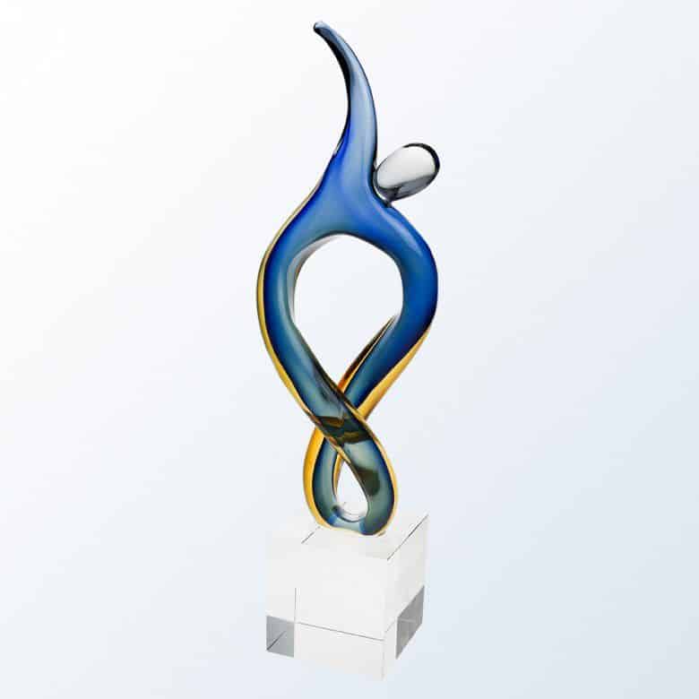 An art glass award featuring a contemporary person reaching for the sky. It has blue and yellow colors mixed in throughout. It's mounted on a clear glass base.