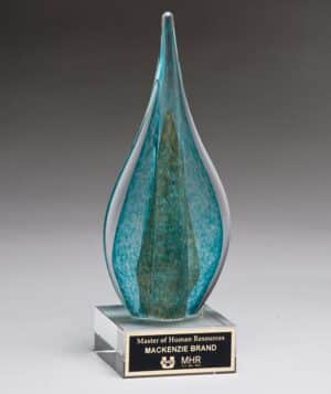 A glass piece in the shape of a flame that has teal & green colors on the inside, along with gold metallic accent as well. It's mounted on a clear glass base which has a black & silver engraving plate.