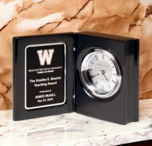 A black book clock on a marble shelf. The book clock features a glossy piano finish, a silver faced clock and a black & silver engraving plate with laser engraving personalization.