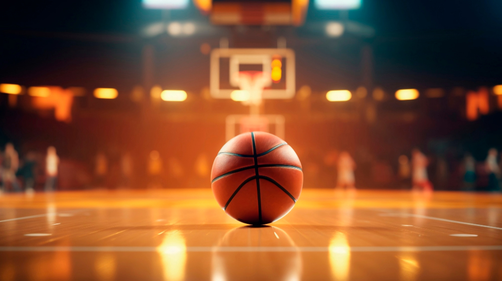 A basketball that is sitting still on a basketball court. In the background is a backboard, hoop & net and on the sides appears to be fans.