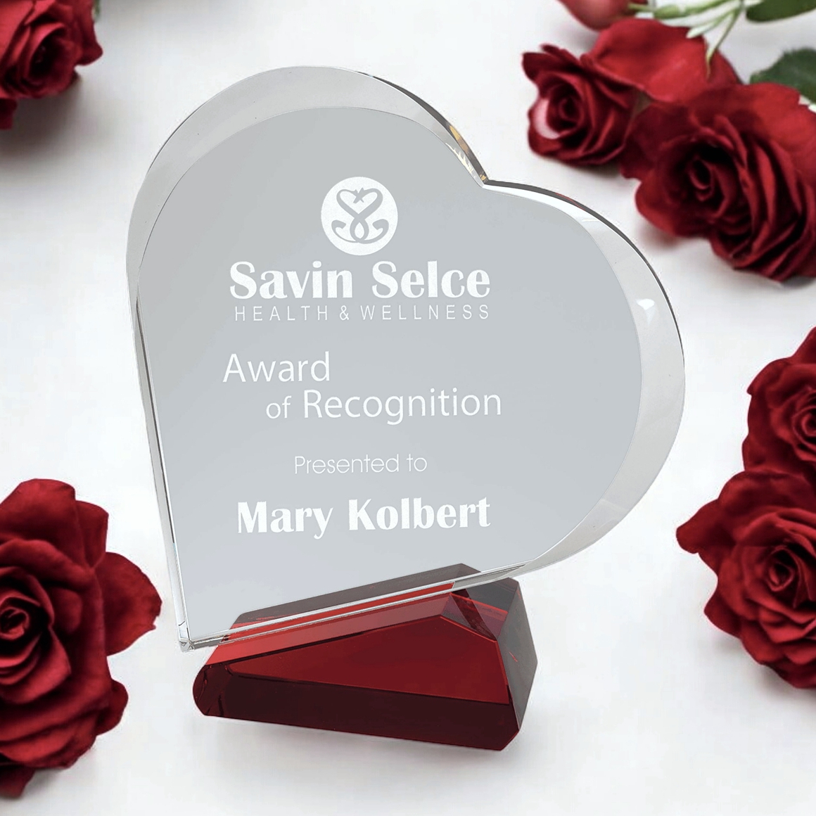 Our Slanted Crystal Heart featuring a clear crystal heart that is personalized with laser engraving. It's mounted on a slanted red crystal base. It's sitting on a white table with red roses surrounding it. This CRY23001RDM is 7" x 7.75" in size, it weighs 4 lbs, comes in a deluxe gift box & includes free engraving!