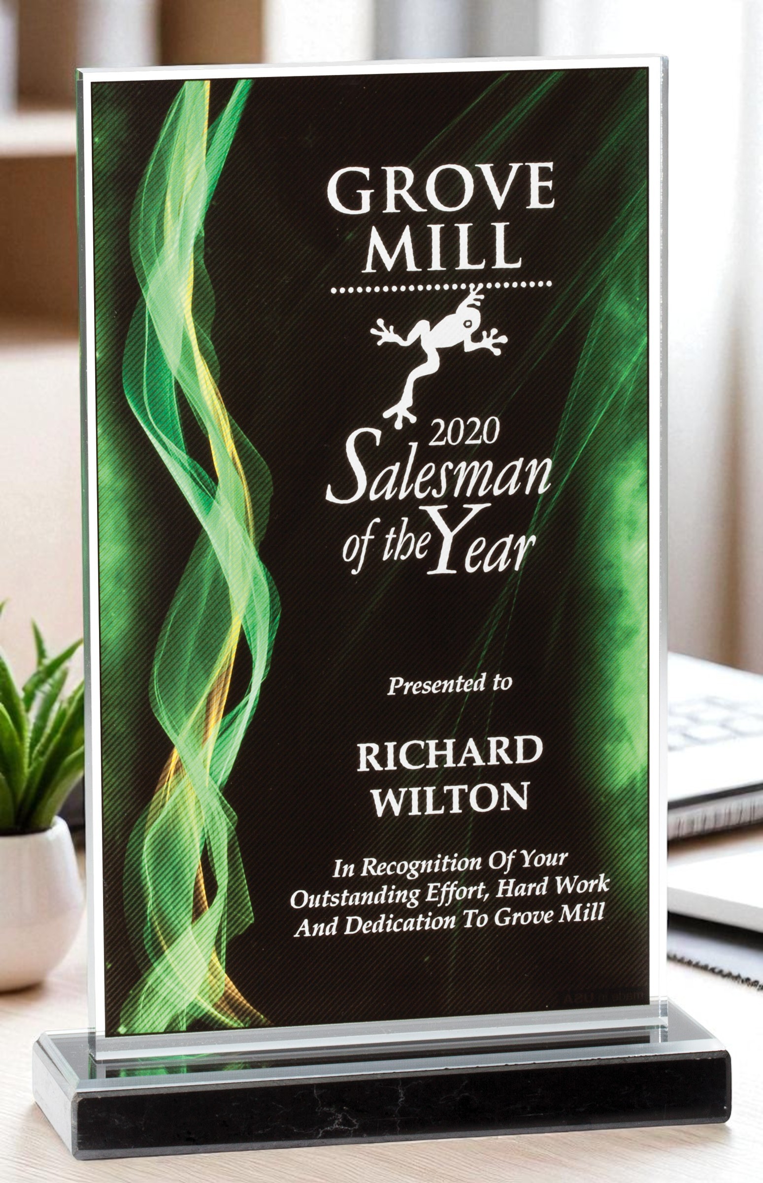 Our Green Illusion Acrylic Award sitting on a desk with a small plant to the left. This award features a rectangle piece of acrylic with a green, yellow & black contemporary design. It's laser engraved for it's recipient who won the Salesman of the Year. This DT1620-G acrylic award is 7" x 11" in size & weighs 3.3 lbs.
