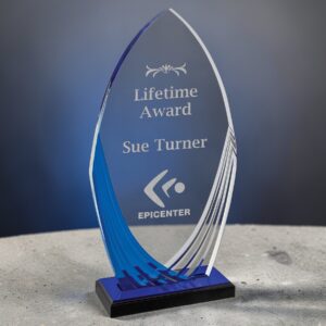 Our Blue Tidal Acrylic Award sitting on a white marble table with a gray background. This is the A7222, which is the 9.75" size. It features a clear acrylic spear with a middle area for laser engraving and blue accents on the left. It's mounted on a black base with a blue mirror top for added blue highlights.