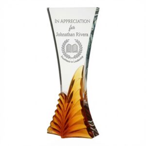 Our Flourish Amber Crystal Award with laser-engraved text in the upper clear section reading "IN APPRECIATION for Johnathan Rivera" above a circular logo of an open book within a laurel wreath and the phrase "Excellence in Leadership". The award has a tall, gracefully curved clear crystal body and an amber-orange fan-shaped base, displayed on a clean white background.