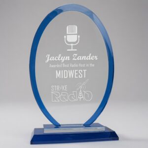 9 inch Oval Blue Regal Glass Award with custom laser engraving. Features a blue border, microphone graphic, and text honoring Jaclyn Zander as Best Radio Host in the Midwest from Strike Radio. Elegant blue glass trophy on a matching base, shown against a light gray background.
